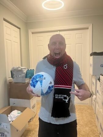 A photo of a man excitedly holding a soccer ball, and pointing at his knit 'Play for Blake' scarf.
